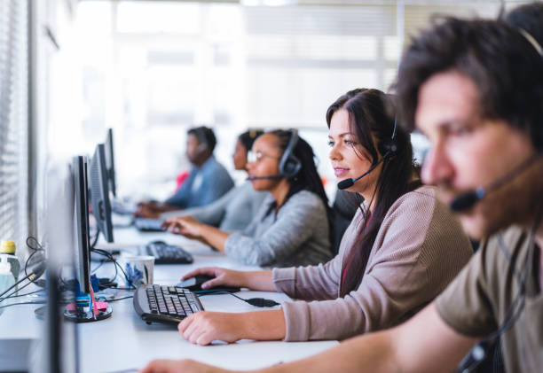 Young businesswoman wearing headset while using computer. Row of operators are sitting at desk. They are working in call center.